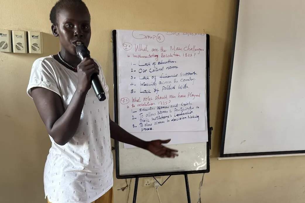 Person standing indoors holding a microphone and pointing toward a flip chart with handwritten notes about challenges in implementing Resolution 1325 and roles played. A blank whiteboard is positioned beside the flip chart.