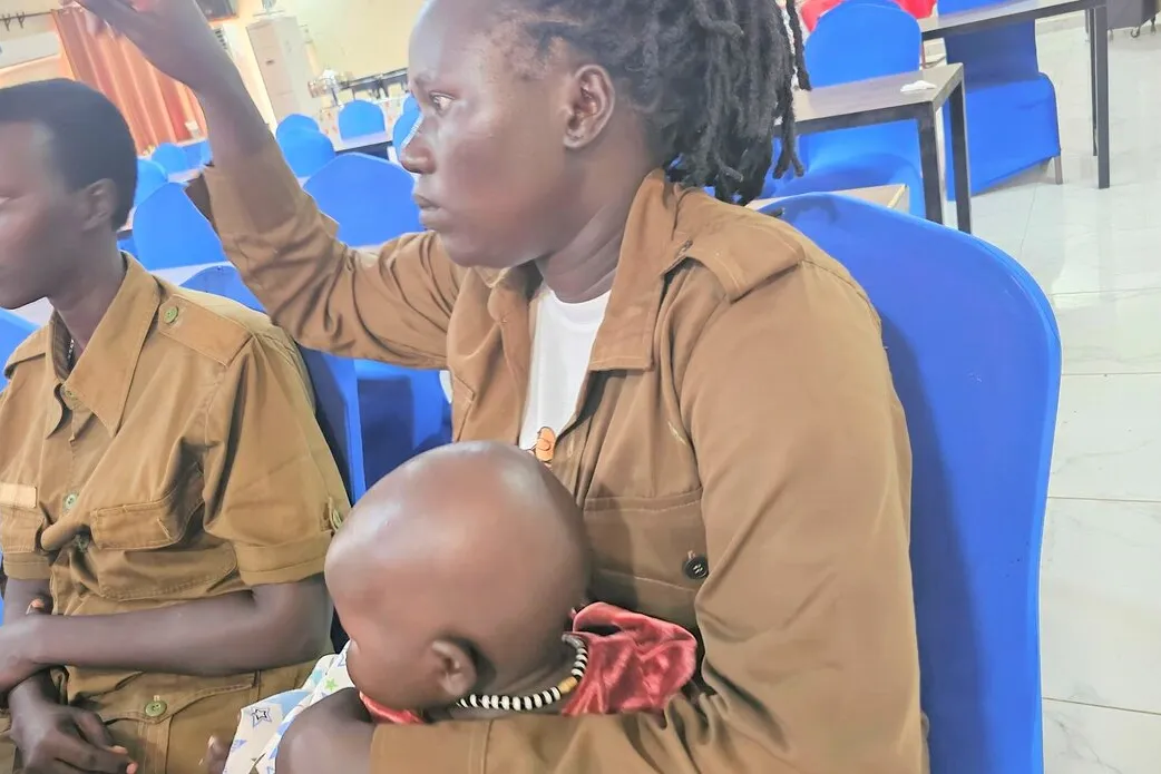Two people in brown uniforms sit on blue chairs indoors, one holding a baby dressed in red, with tables and more chairs visible in the background