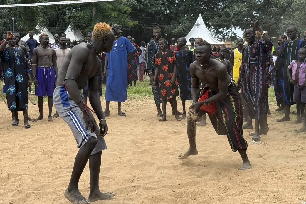 Two individuals engaged in a traditional wrestling match on sandy ground, surrounded by a crowd wearing colorful patterned clothing, with white tents and trees in the background.