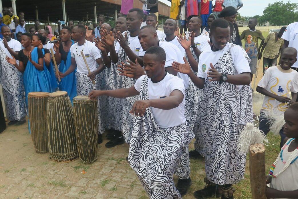Group of people wearing white shirts and patterned wraps performing a traditional dance outdoors, with drums placed in front and others clapping along.