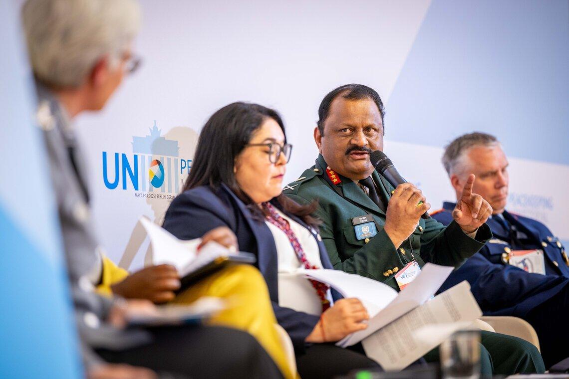 two men and two women panelists on a stage. A man in a military uniform is holding a microphone.