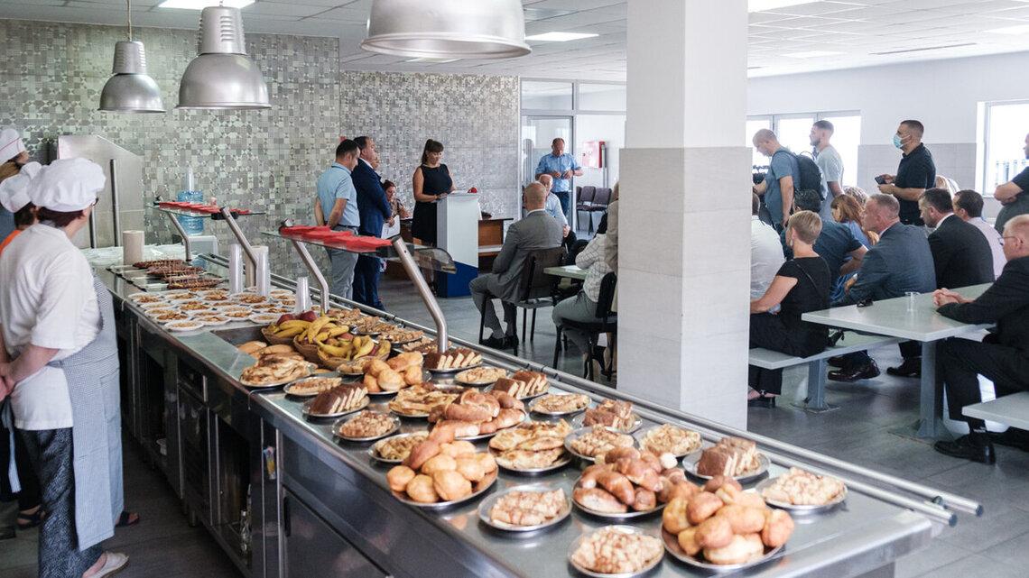 People gather in a cafeteria-style setting with trays of baked goods and fruit neatly arranged on a serving counter, while chefs stand nearby and a woman addresses the seated audience.