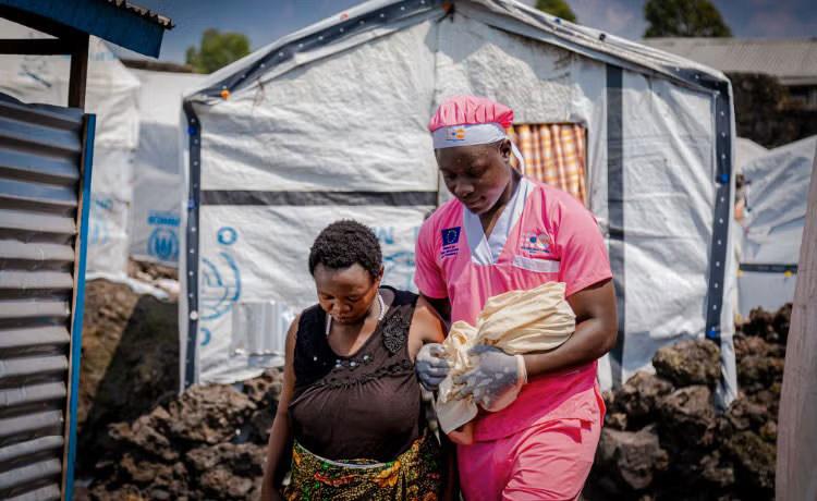 A healthcare worker in pink uniform assists a person near tents marked with UNHCR and EU logos.
